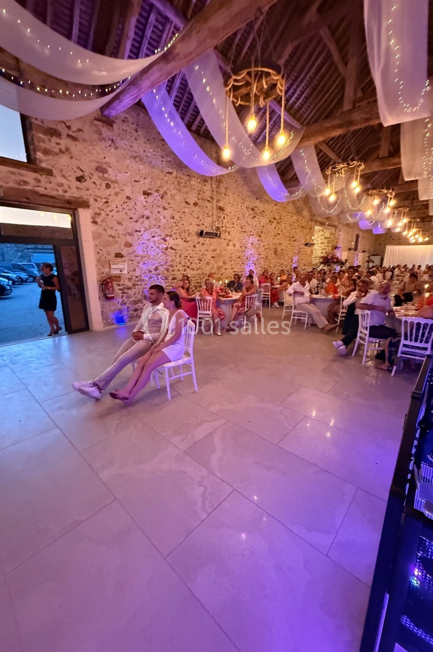 Salle de réception décorée avec des guirlandes lumineuses, des invités assis à des tables et un couple au premier plan.