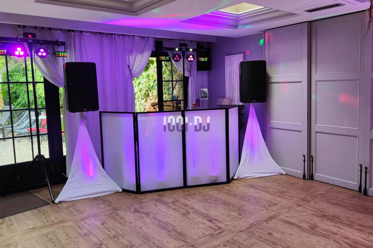 Poste de DJ installé dans une salle avec éclairage coloré, rideaux blancs et parquet au sol.