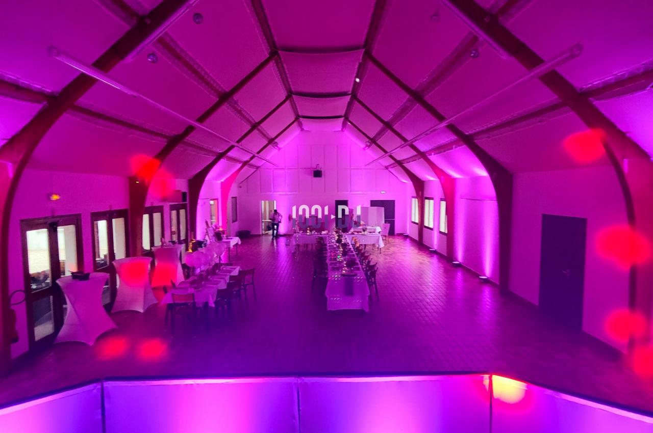 Salle de réception spacieuse éclairée par des lumières roses et violettes, avec tables dressées et décorations.
