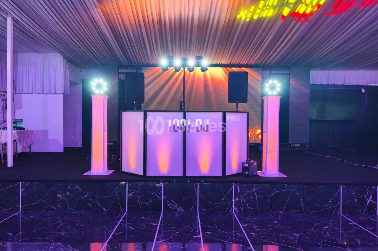 Console de DJ éclairée en blanc et violet, entourée de haut-parleurs et de colonnes lumineuses dans une salle de réception.