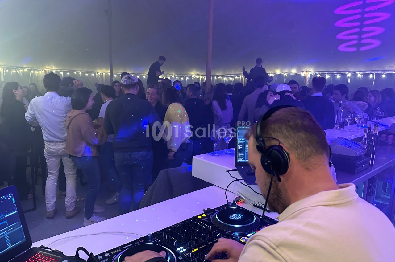Un DJ mixe sur une table de mixage devant une foule dansante sous une tente éclairée par des guirlandes lumineuses.