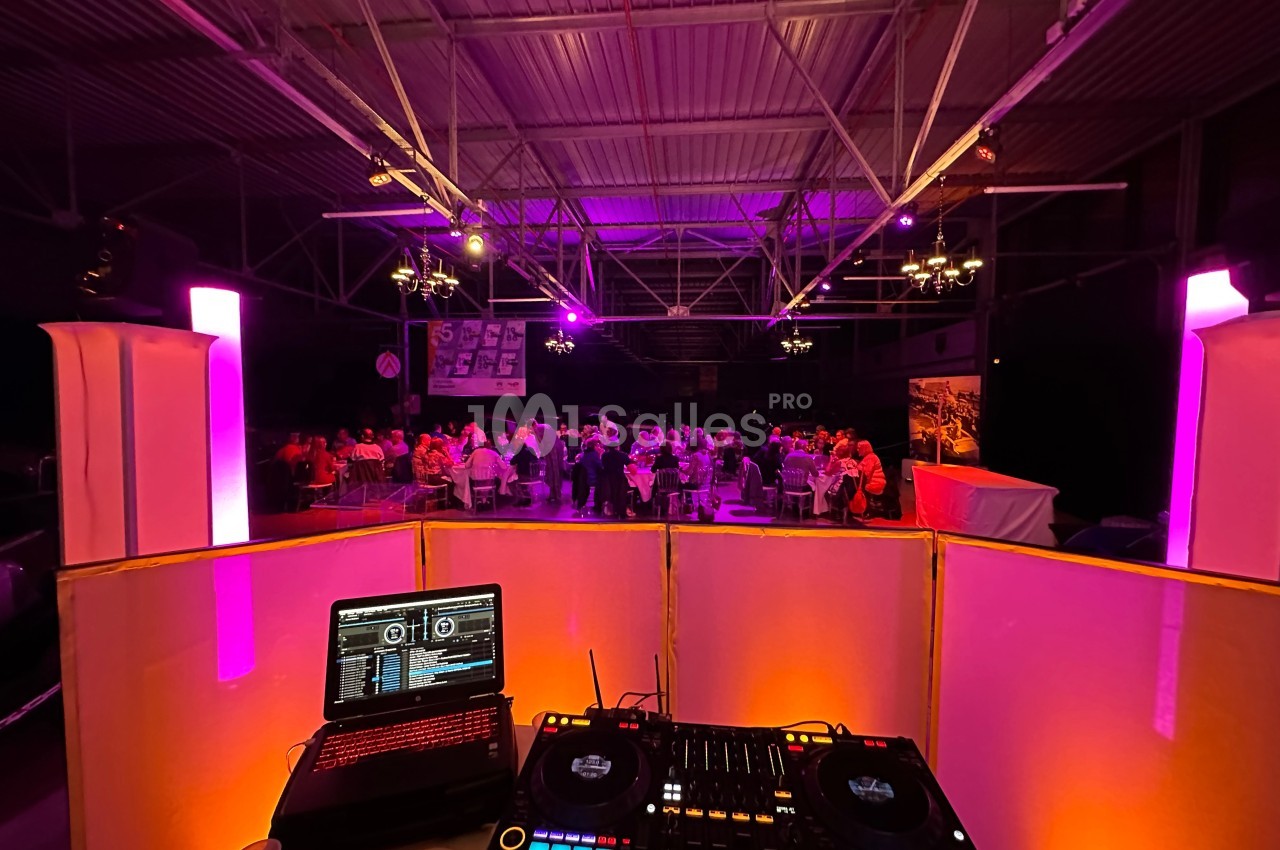 Vue depuis une cabine de DJ avec platines et ordinateur, éclairage coloré et invités assis dans une salle sombre.