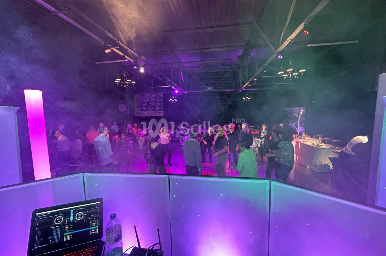 Vue depuis une cabine de DJ sur une piste de danse animée, éclairée par des lumières colorées et entourée de fumée légère.