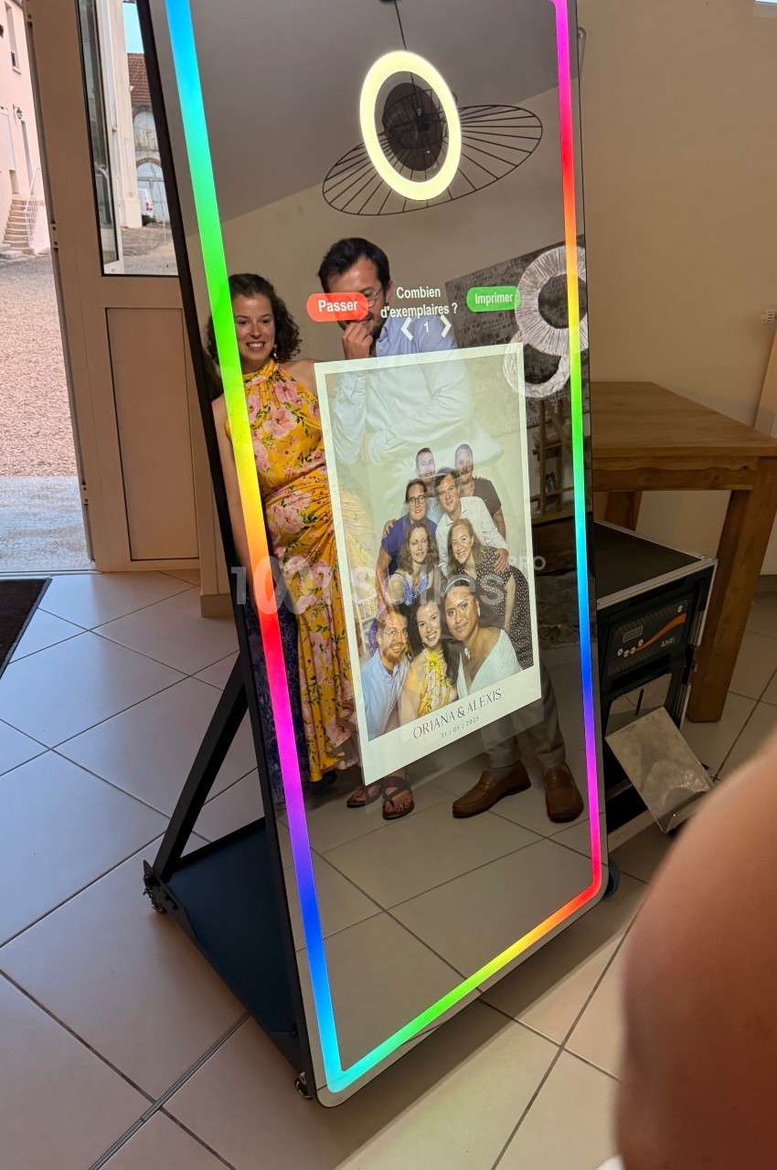 Un miroir photo interactif reflète un couple souriant et affiche une photo imprimée de groupe en bas.