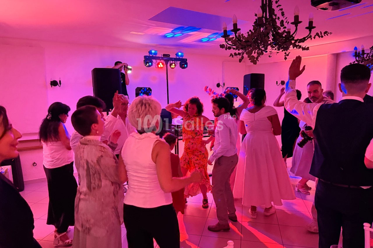 Des personnes dansent dans une salle éclairée par des lumières colorées, avec un DJ et des chandeliers au plafond.