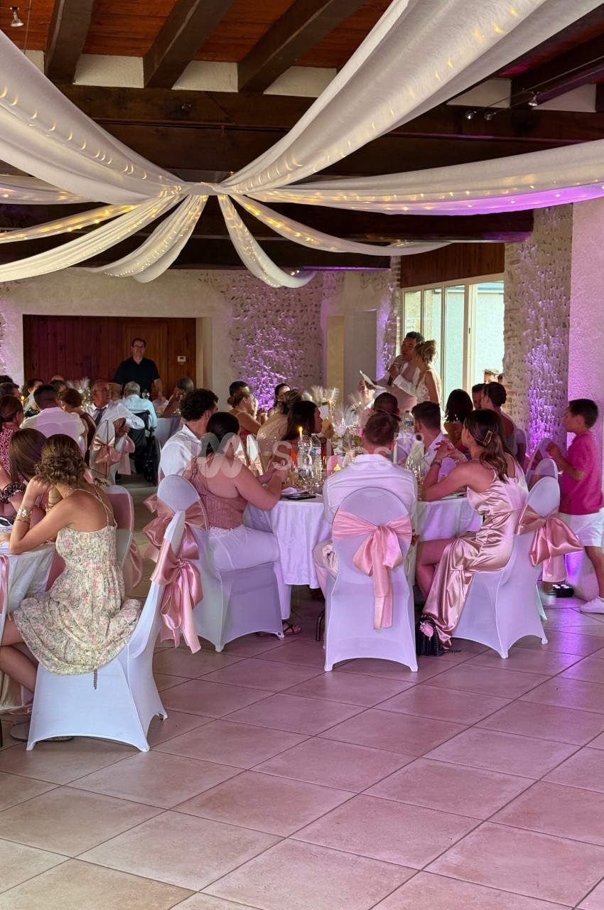 Salle de réception décorée avec des nappes blanches et roses, des invités assis autour de tables lors d'un événement festif.