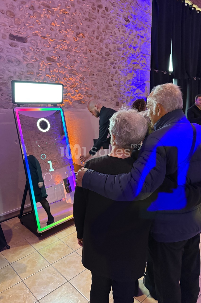 Un couple âgé observe un miroir interactif lumineux dans une salle aux murs en pierre, entouré de plusieurs personnes.