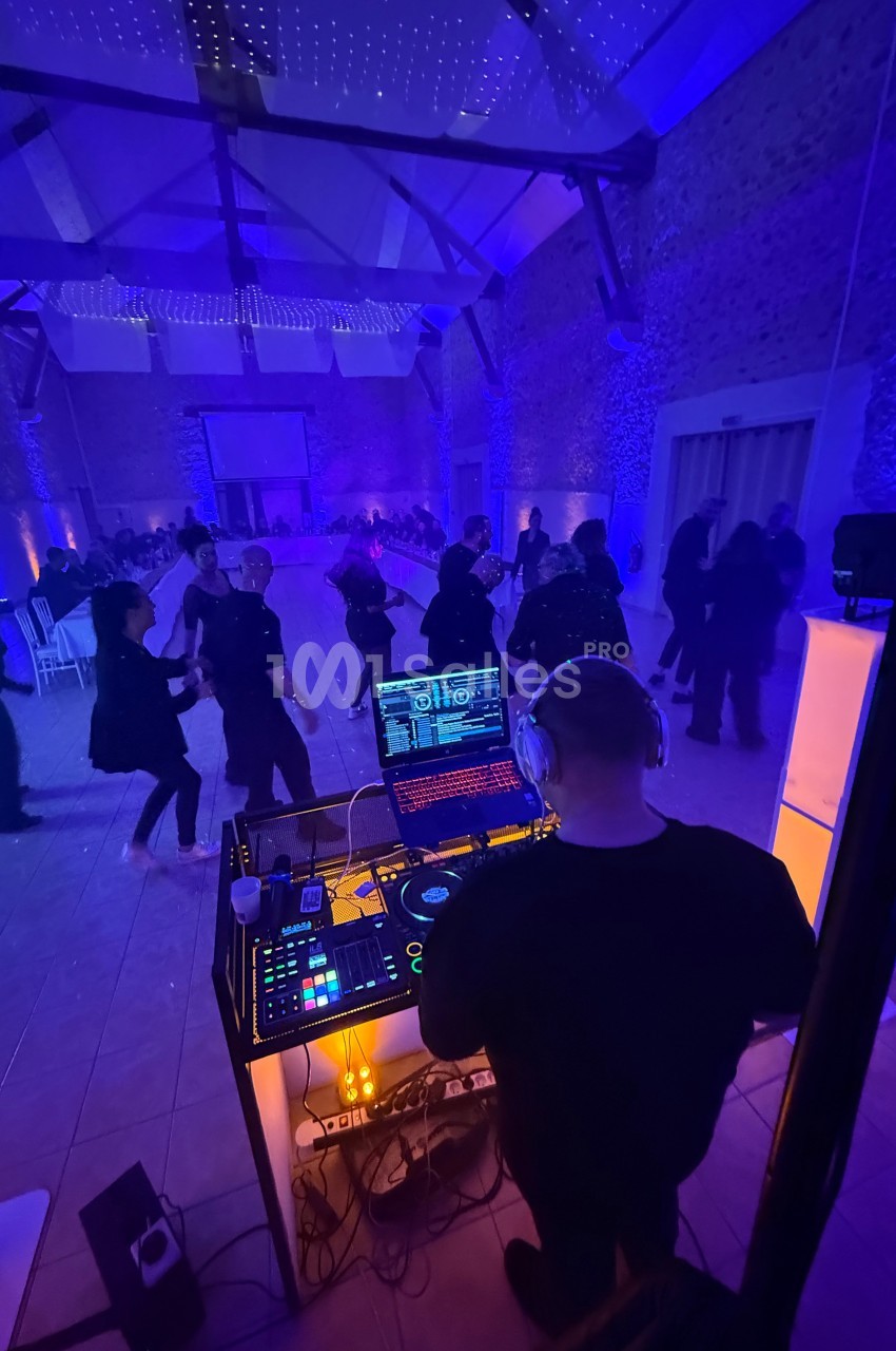 Un DJ mixe devant une piste de danse où des personnes profitent d'une ambiance lumineuse violette dans une salle en pierre.