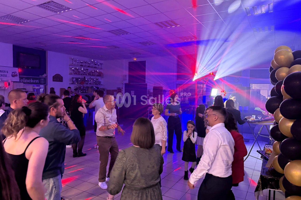 Des personnes dansent et discutent dans une salle éclairée par des lumières colorées, décorée de ballons noirs et dorés.