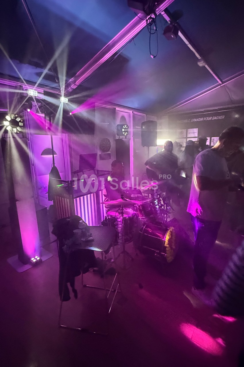 Un batteur joue sur scène dans une ambiance lumineuse violette et tamisée, entouré de spectateurs.