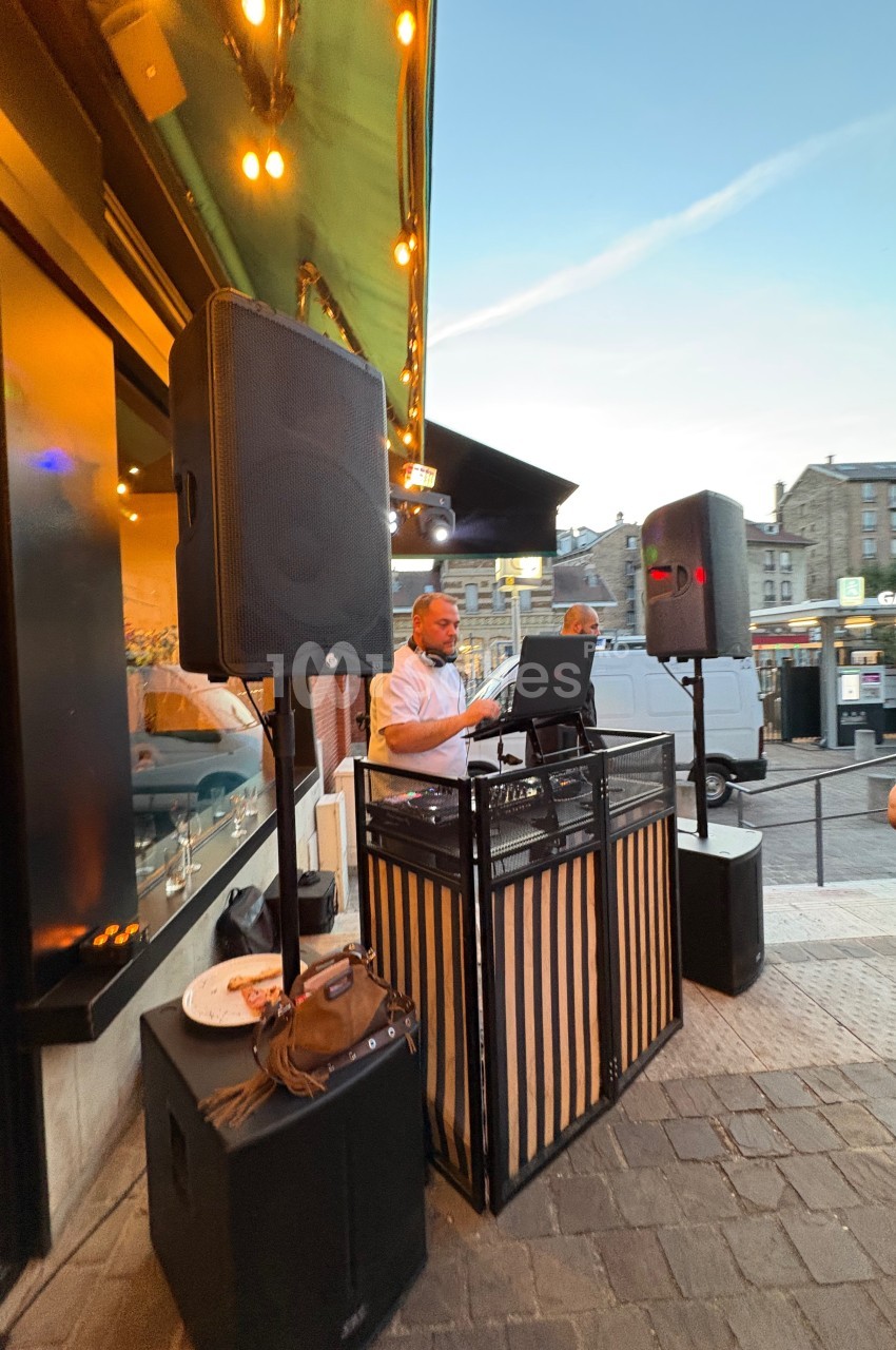 Un DJ mixe en extérieur devant un restaurant, entouré de haut-parleurs, au crépuscule dans une rue animée.