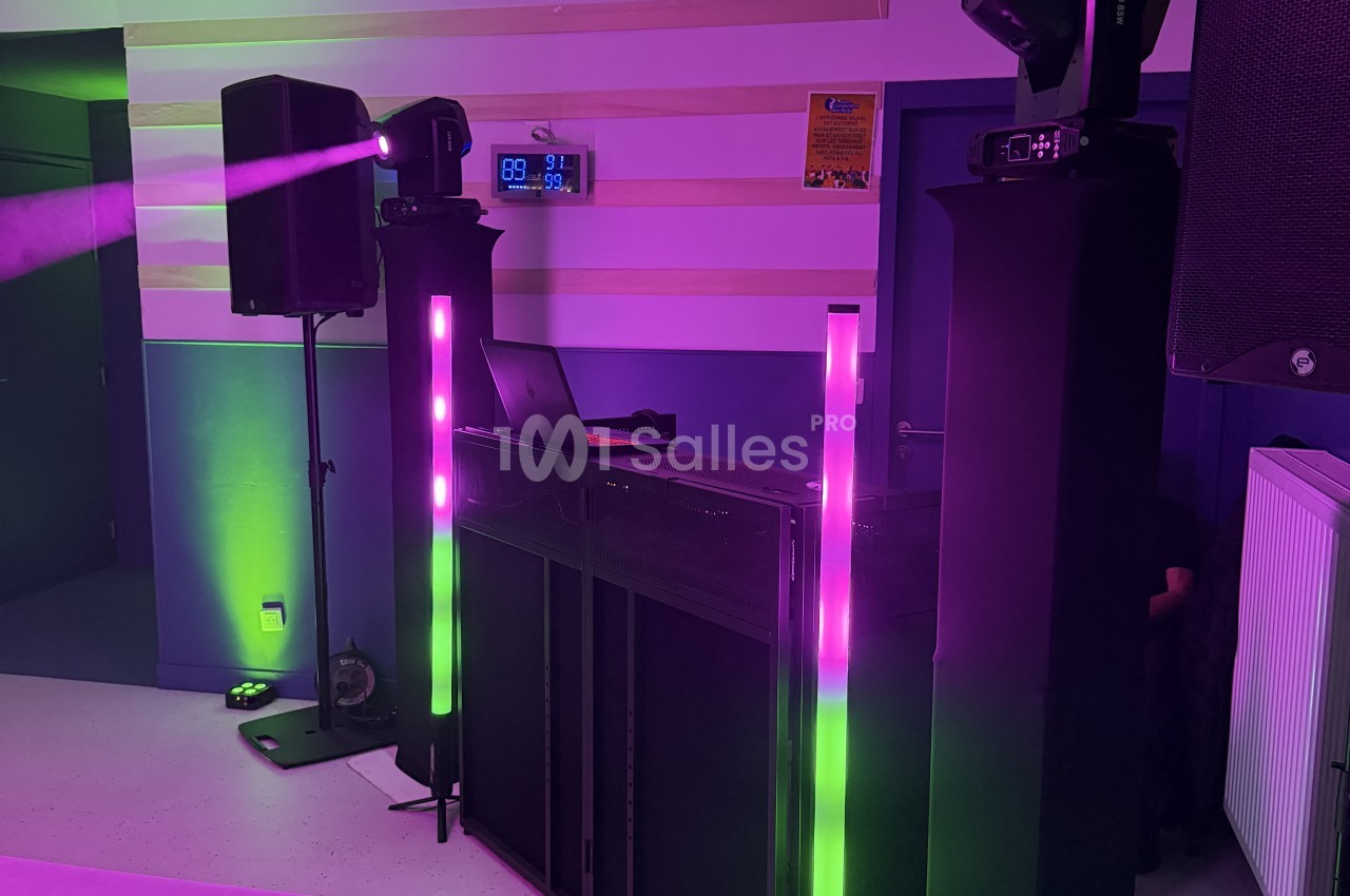 Installation de DJ dans une pièce aux murs violets et verts, avec enceintes sur pied, jeux de lumières colorées, tubes LED verticaux et ordinateur portable posé sur une table. Let me check length: roughly 165 characters, too long. Need ≤125. Installation
