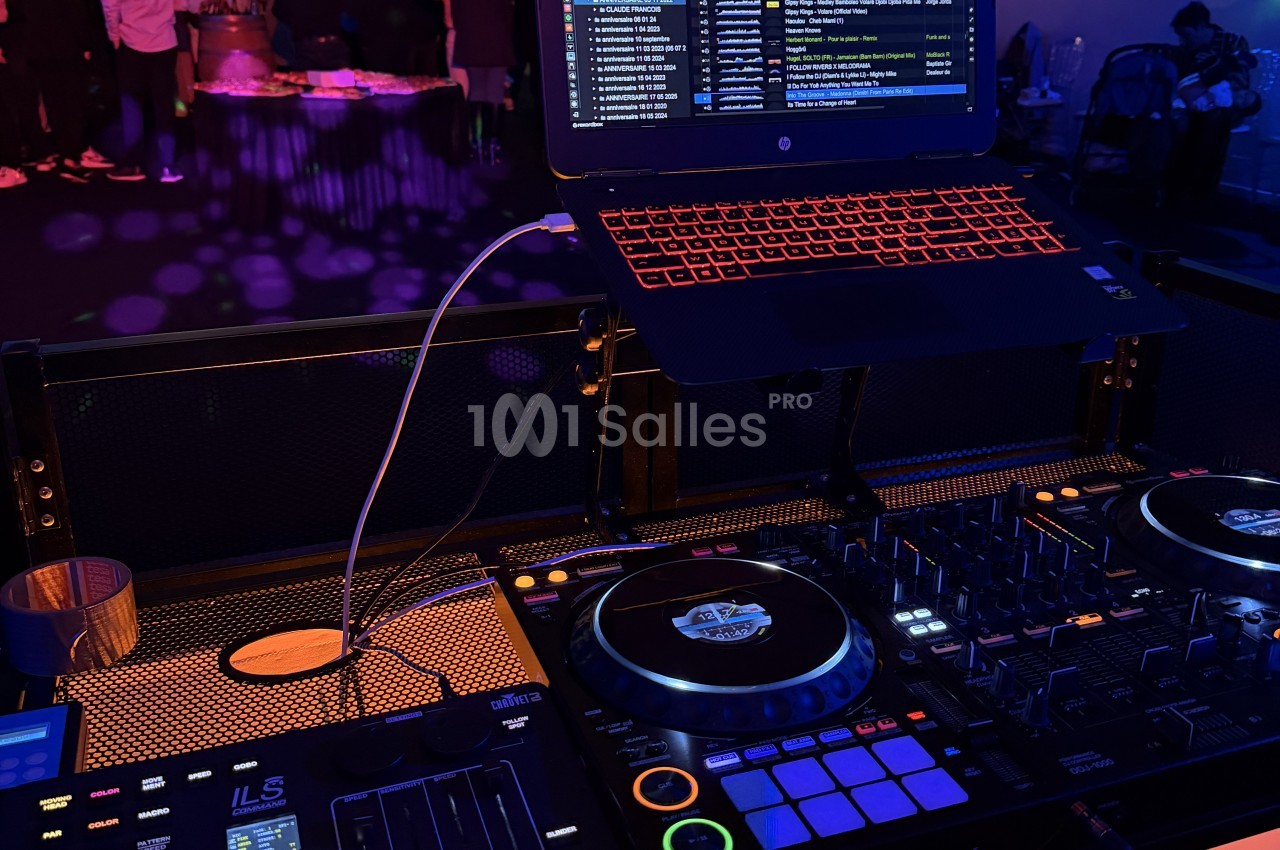 Poste de DJ en soirée : contrôleur à platines et table de mixage sous éclairage violet et orangé, ordinateur portable posé sur un support, piste de danse floue à l'arrière-plan.