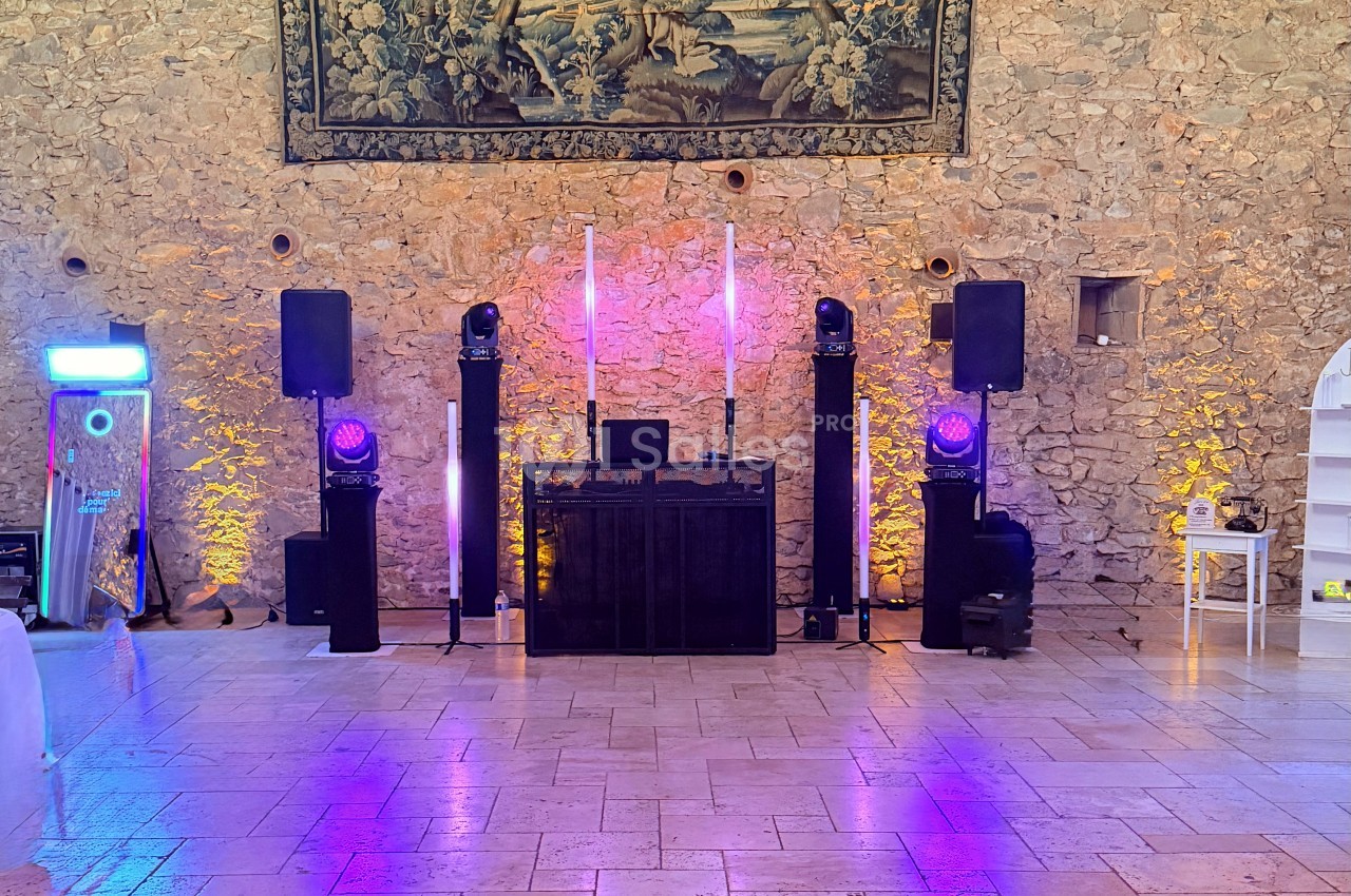 Installation de DJ dans une salle aux murs de pierre apparente, avec enceintes, projecteurs à tête mobile violets, colonnes lumineuses et une cabine de platines sur un sol carrelé. Let me verify length: roughly 170 characters, too long. Need ≤125.