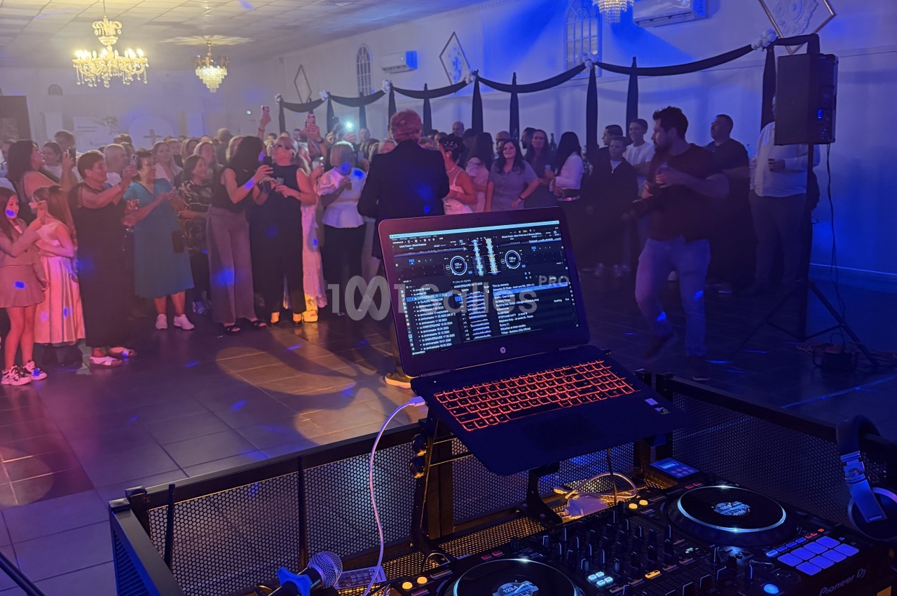 Poste de DJ avec ordinateur portable et platines devant une piste de danse où des invités se rassemblent sous des lustres et un éclairage bleu.