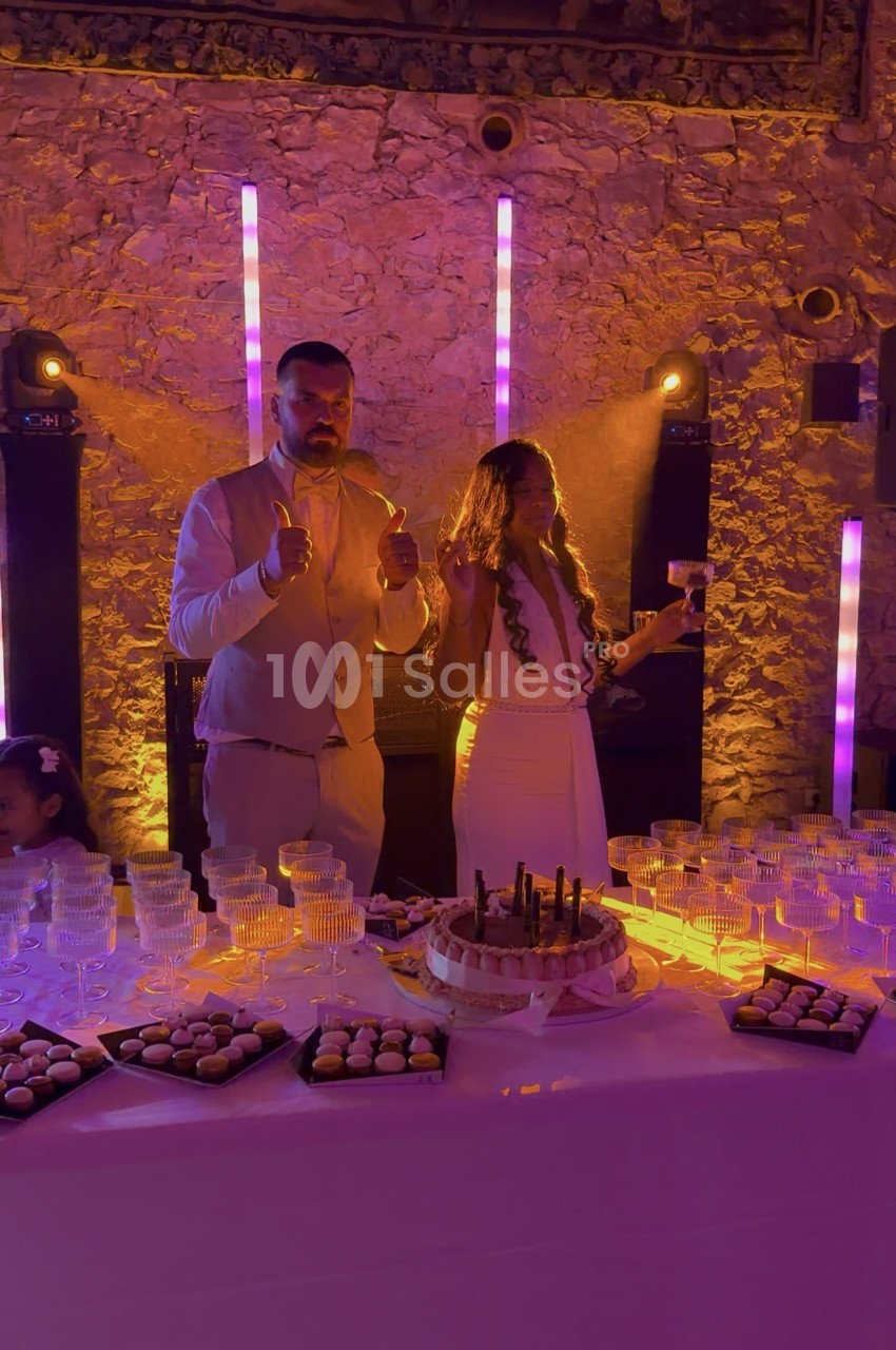 Deux personnes debout derrière une table de buffet en soirée, un pouce levé, devant un mur de pierre éclairé en orangé et des barres lumineuses violettes, avec gâteau et petits fours. Count: roughly 170 characters? Let's shorten to ≤125. Deux personnes