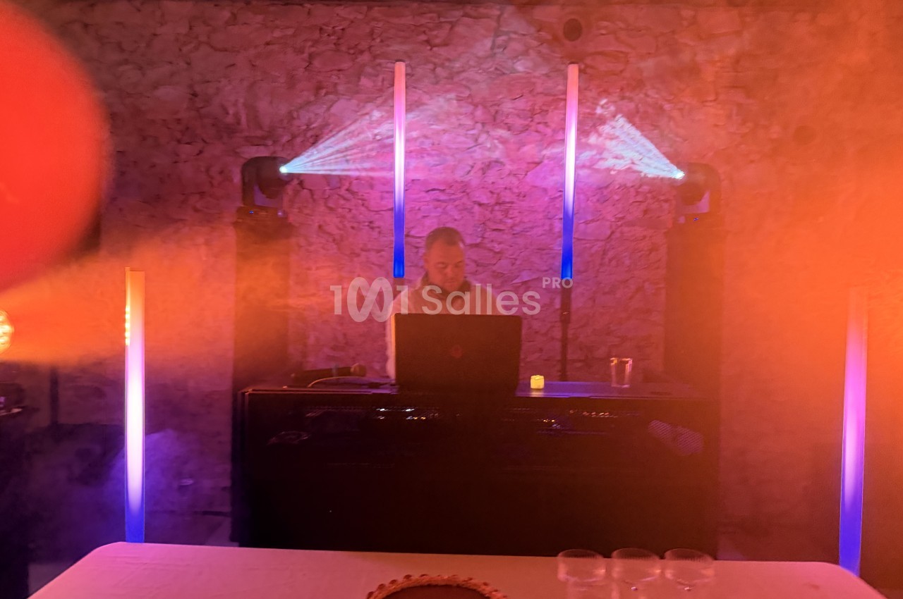 Un DJ derrière sa console, un ordinateur portable posé devant lui, dans une pièce aux murs en pierre éclairée par des jeux de lumières violettes et orangées, avec des projecteurs et des barres lumineuses verticales. Let me check length: roughly 215