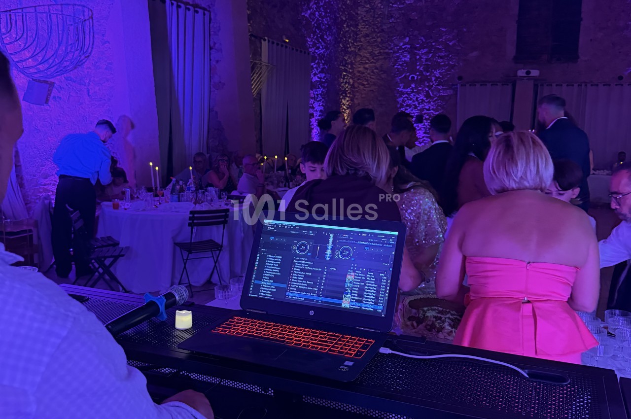 Un DJ derrière une table de mixage avec ordinateur portable et micro, face à une salle de réception éclairée en violet où des convives sont attablés.