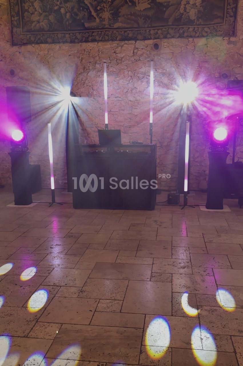 Installation de DJ dans une salle à murs en pierre apparente, avec projecteurs violets, barres lumineuses verticales et jeux de lumière sur le sol carrelé.