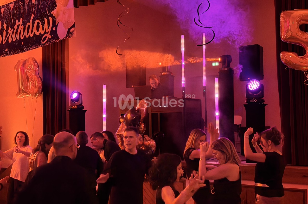 Soirée d'anniversaire en intérieur : des invités dansent sous un éclairage violet et orangé, un DJ est installé derrière sa console au fond, avec ballons dorés et enceintes sur les côtés. Let me check length: roughly 165 characters, too long. Need ≤125.