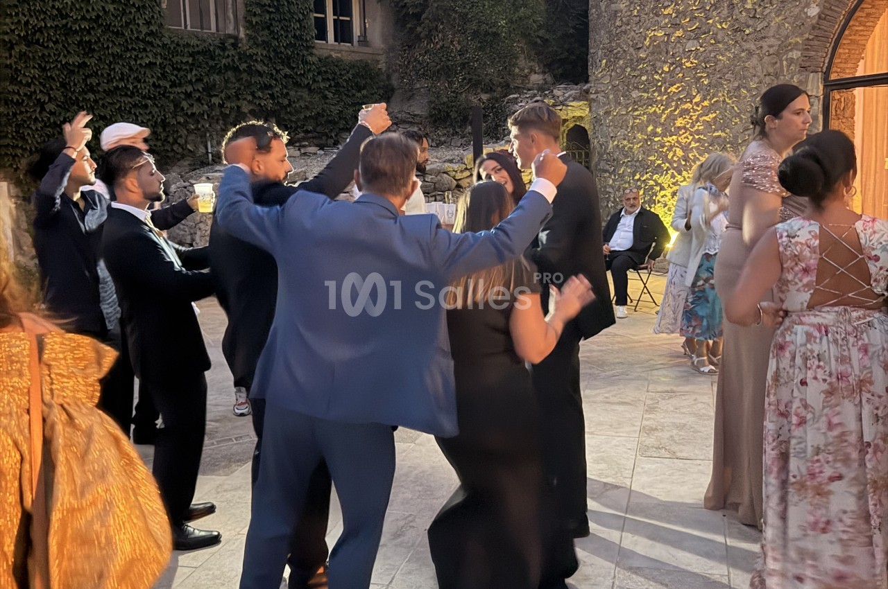 Des convives dansent en groupe sur une terrasse pavée en soirée, certains tenant des gobelets, près d'un mur de pierre couvert de lierre.