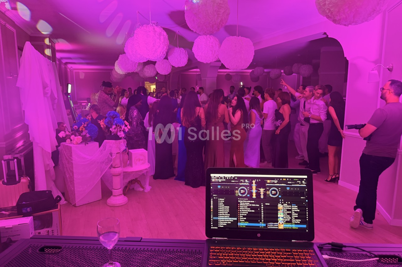 Soirée dansante dans une salle éclairée en violet, avec des convives sur la piste sous des boules blanches suspendues, et un ordinateur de DJ au premier plan.