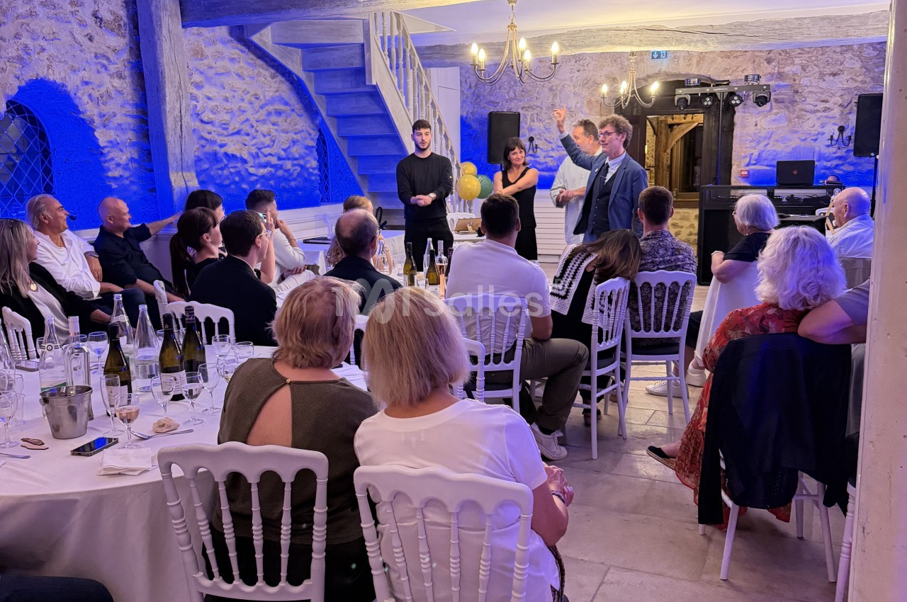Soirée dans une salle aux murs de pierre éclairés en bleu, des convives attablés écoutent un homme qui prend la parole debout devant un escalier. Let me check length: roughly 150 characters, too long. Need ≤125. Shorter: "Convives attablés dans une salle