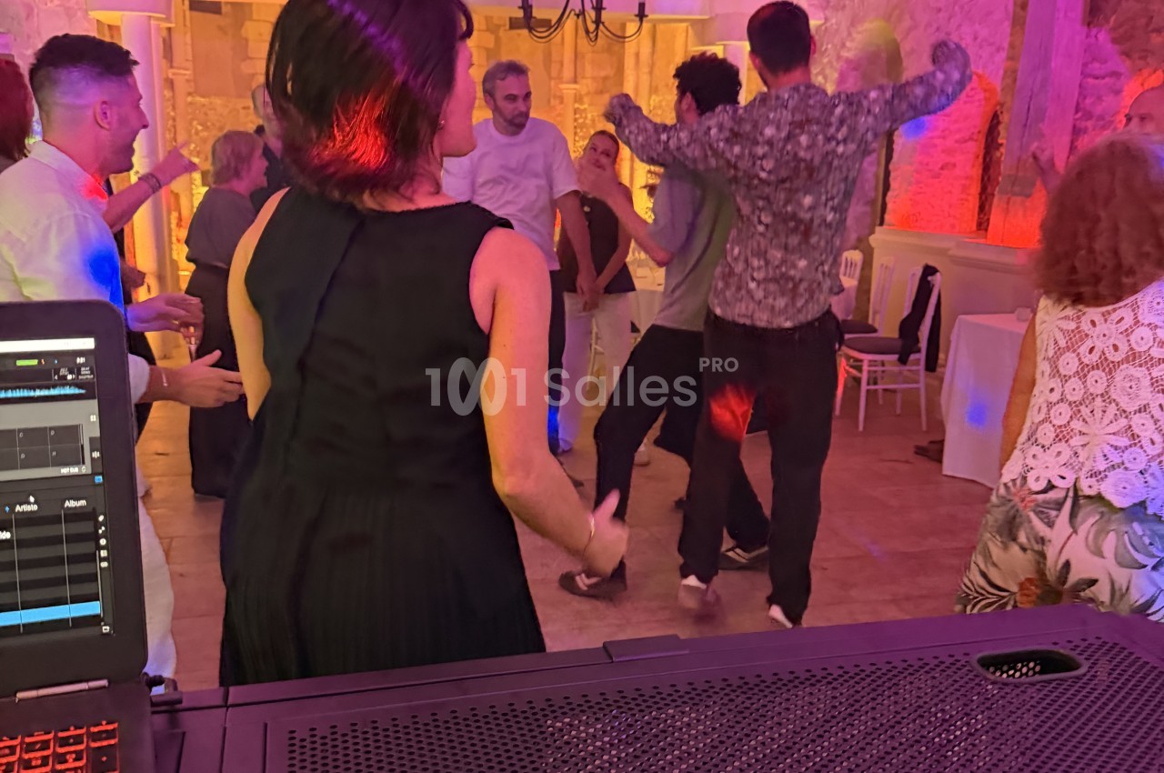 Un DJ derrière sa table de mixage règle la musique tandis que des convives dansent sur une piste éclairée aux tons violets et rouges. Let me check length: roughly 130 characters? Count: "Un DJ derrière sa table de mixage règle la musique tandis que des