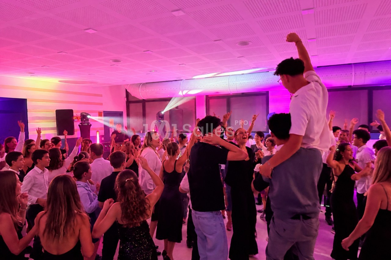 Des jeunes dansent en nombre dans une salle éclairée de lumières roses et violettes, sous un plafond acoustique, avec des projecteurs au fond.
