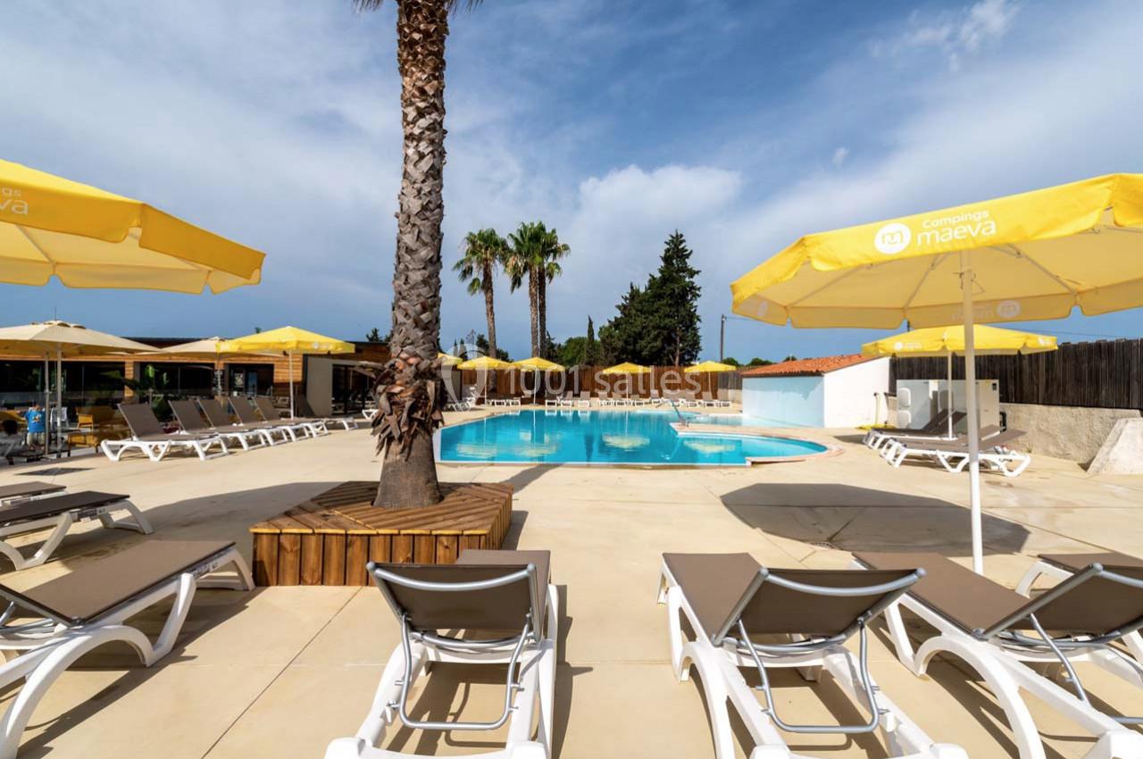 Piscine extérieure entourée de transats, parasols jaunes et palmiers sous un ciel partiellement nuageux.