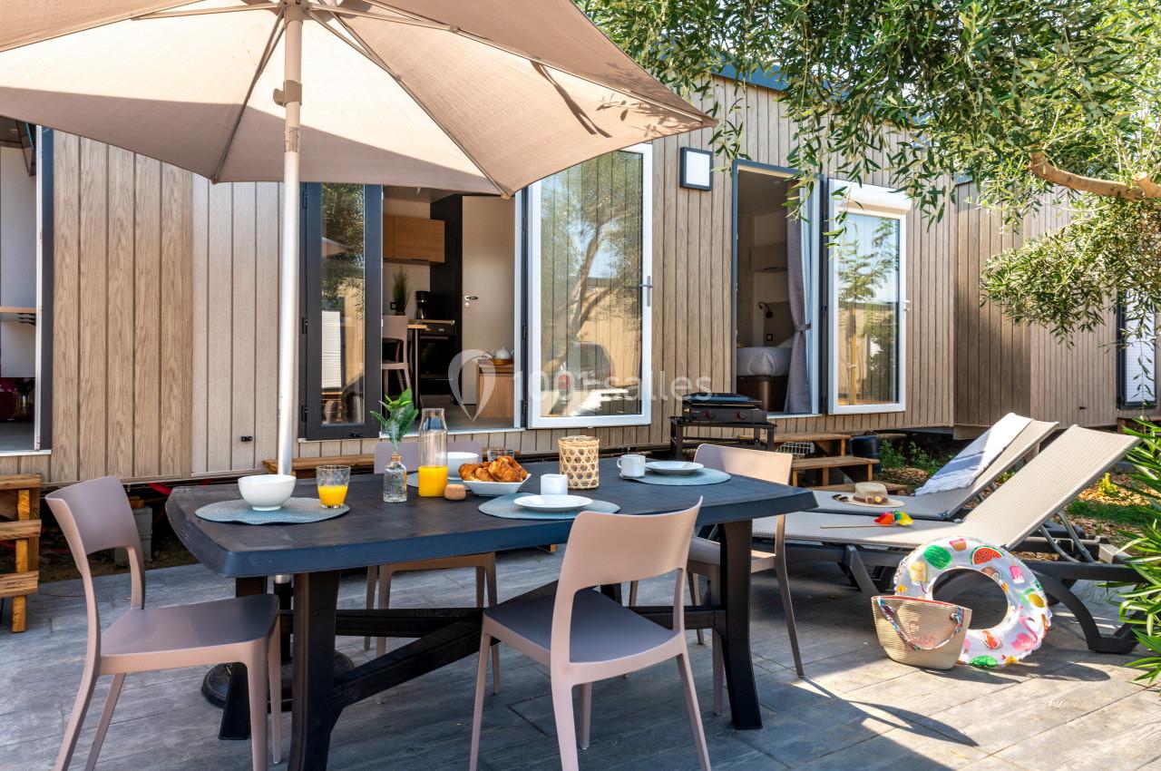Terrasse aménagée avec table dressée pour un repas, chaises, parasol, transat et vue sur un logement moderne en bois.