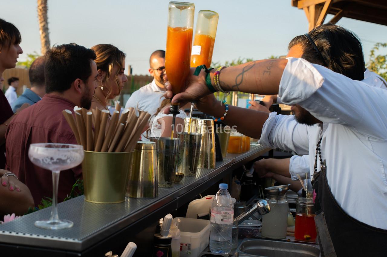 Un barman prépare des cocktails devant un groupe de personnes lors d'un événement en extérieur au coucher du soleil.