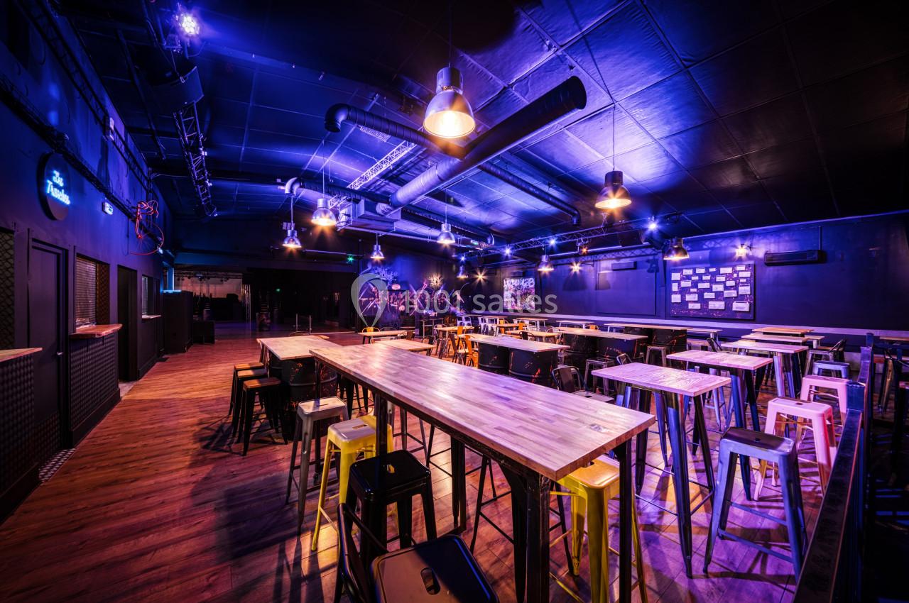 Salle spacieuse avec tables hautes, tabourets colorés et éclairage violet dans une ambiance industrielle.