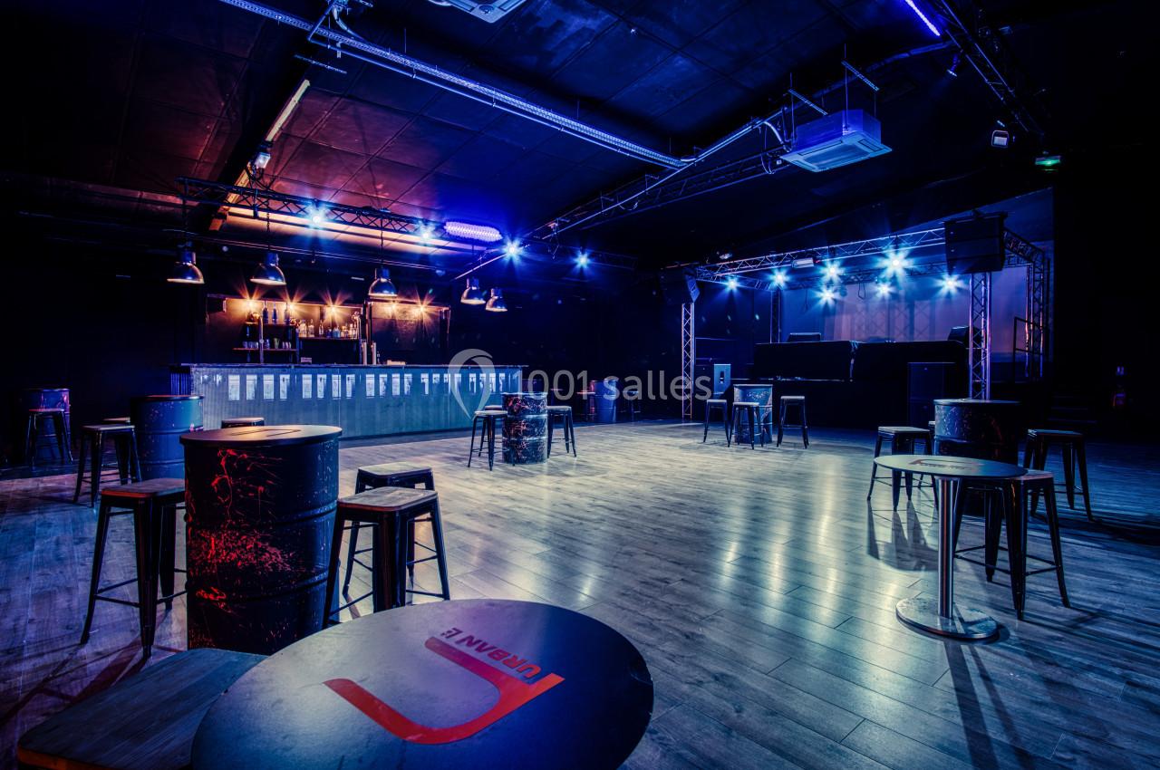 Salle de concert vide avec éclairage bleu, tables hautes et tabourets dispersés, scène et bar en arrière-plan.