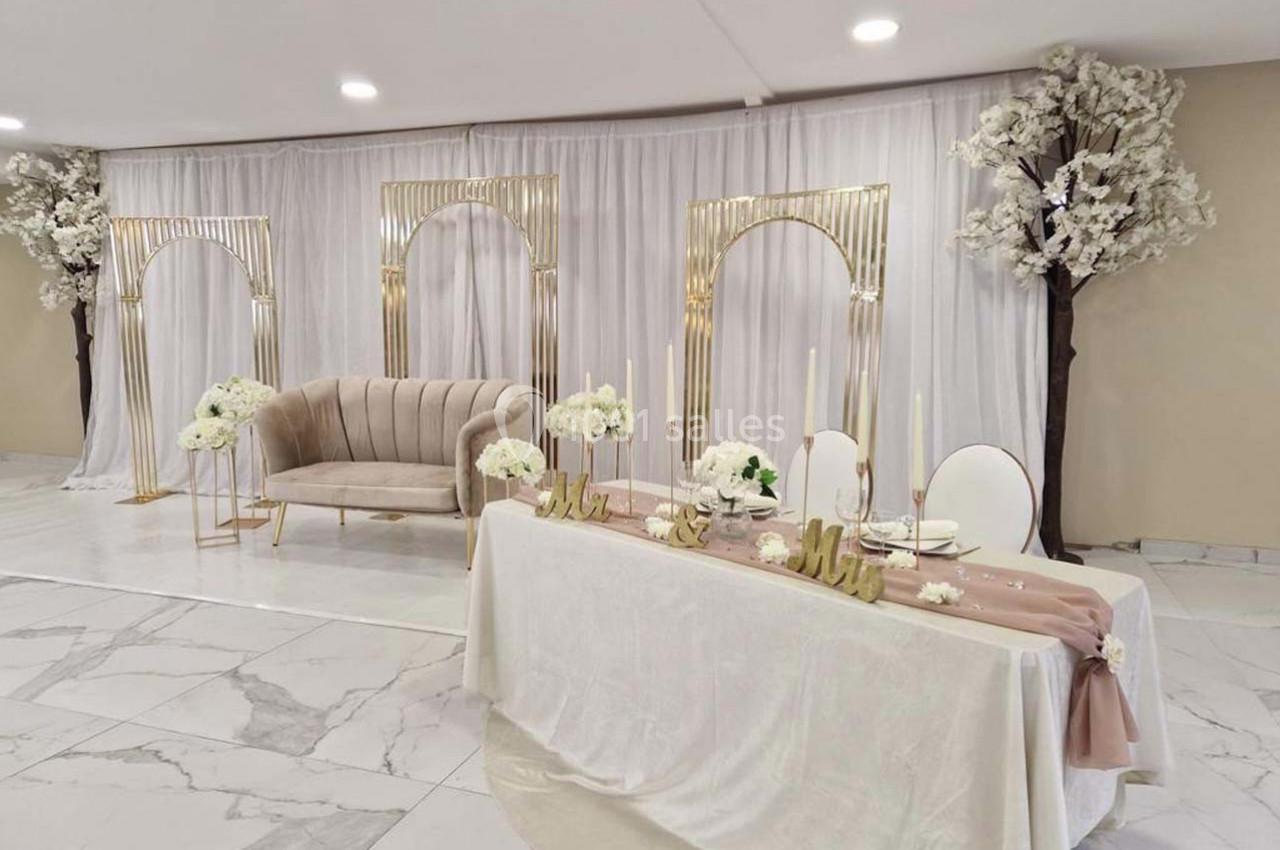 Salle décorée pour un événement avec arche dorée, canapé beige, table ornée de fleurs et chandeliers.