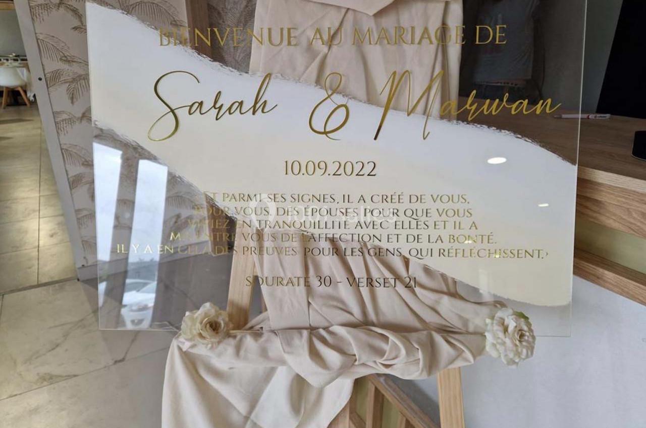 Panneau de mariage transparent avec inscriptions dorées, posé sur un chevalet décoré de tissu et de fleurs.