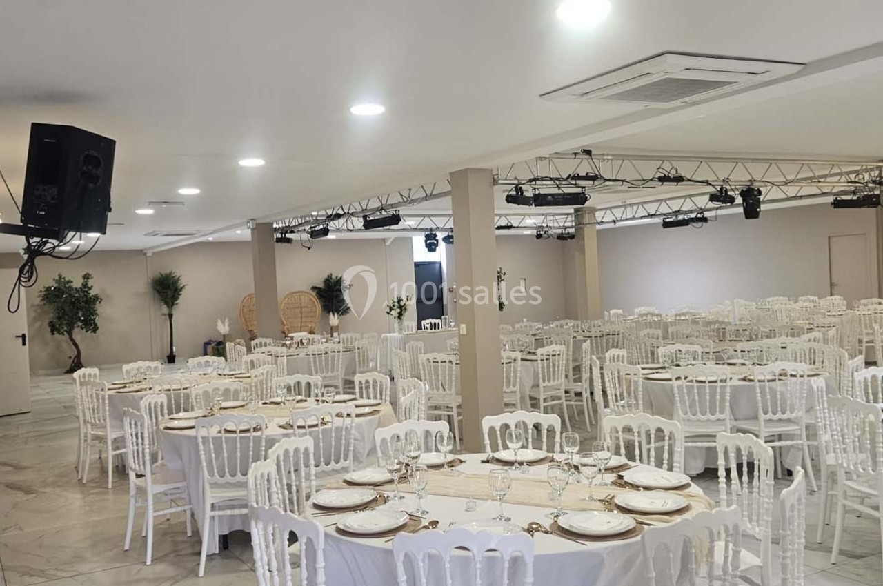 Salle de réception lumineuse avec tables rondes dressées, chaises blanches et décoration sobre.
