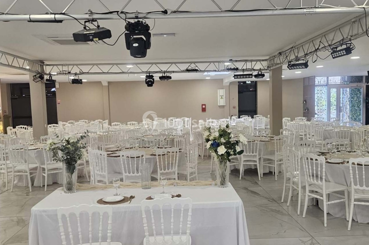Salle de réception lumineuse avec tables rondes dressées, chaises blanches et décorations florales.