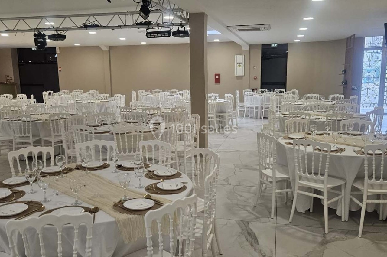 Salle de réception aménagée avec des tables rondes dressées, nappes blanches et chaises en bois blanc.