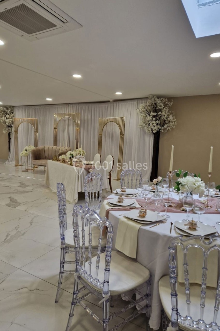 Salle de réception élégante avec tables décorées, chaises transparentes et arrière-plan drapé blanc orné d'arcs dorés.