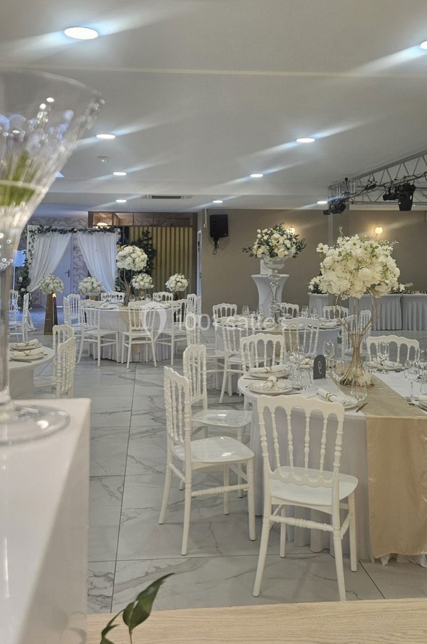 Salle de réception décorée avec des tables dressées, chaises blanches et arrangements floraux blancs.