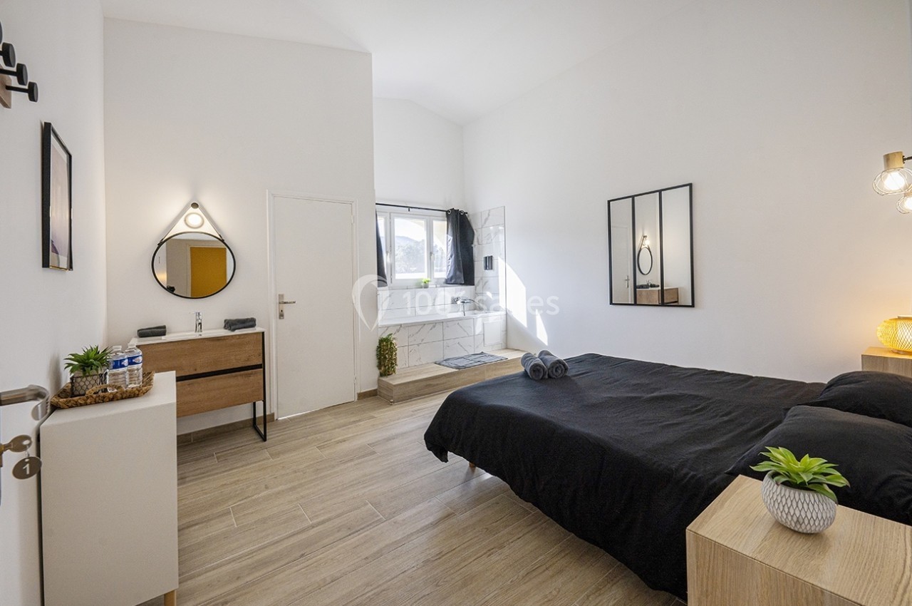 Chambre lumineuse avec lit double, mobilier moderne, miroir rond, salle de bain attenante et grande fenêtre.