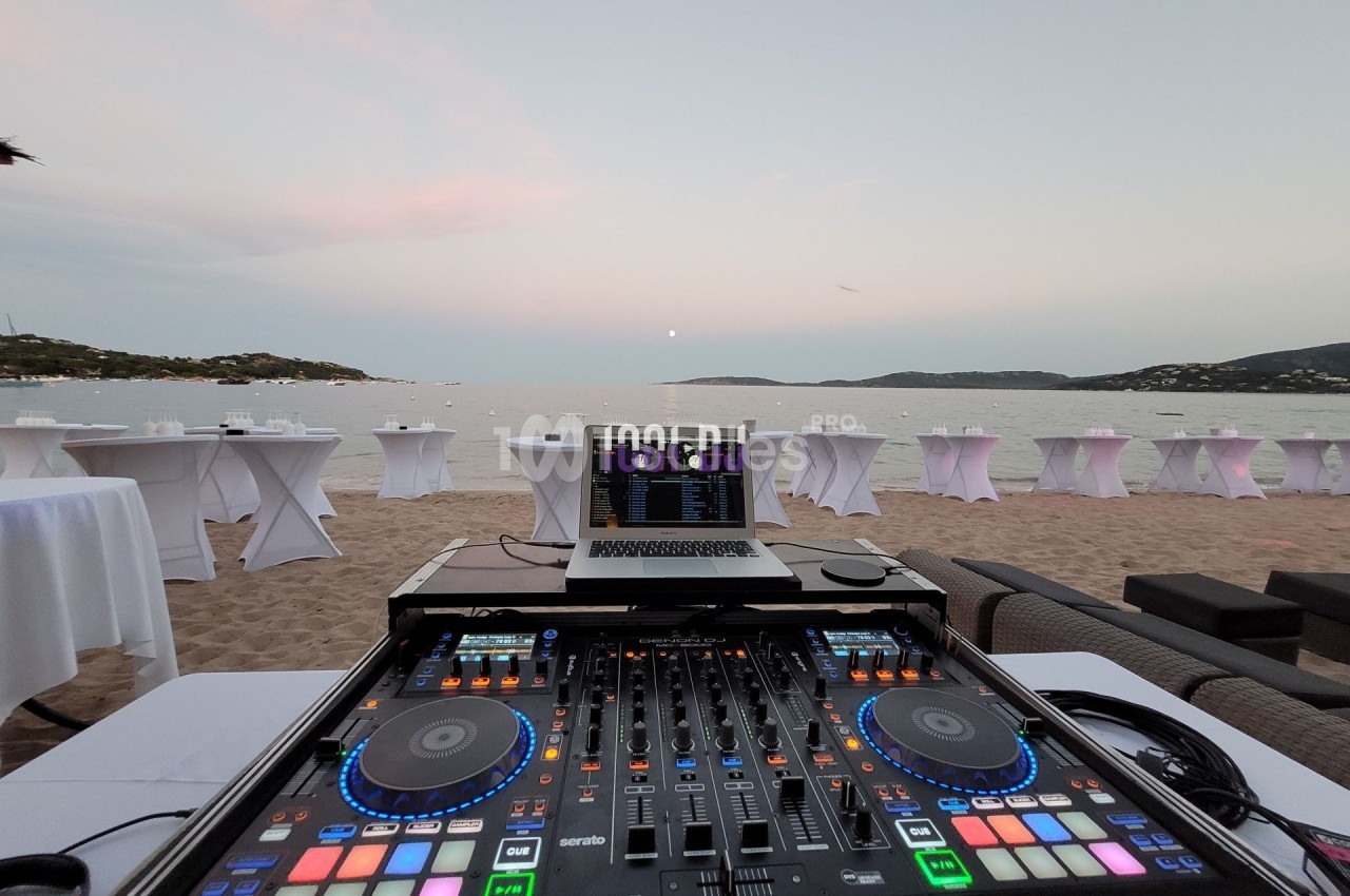 Table de mixage DJ sur une plage au coucher du soleil, avec des tables dressées en arrière-plan près de la mer.