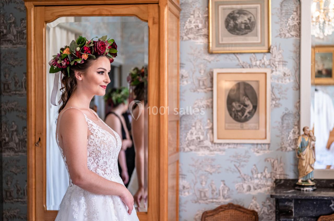 Location salle Malviès (Aude) - Château Guilhem #1 Femme en robe de mariée avec une couronne de fleurs, debout devant un miroir dans une pièce décorée de cadres et papiers…