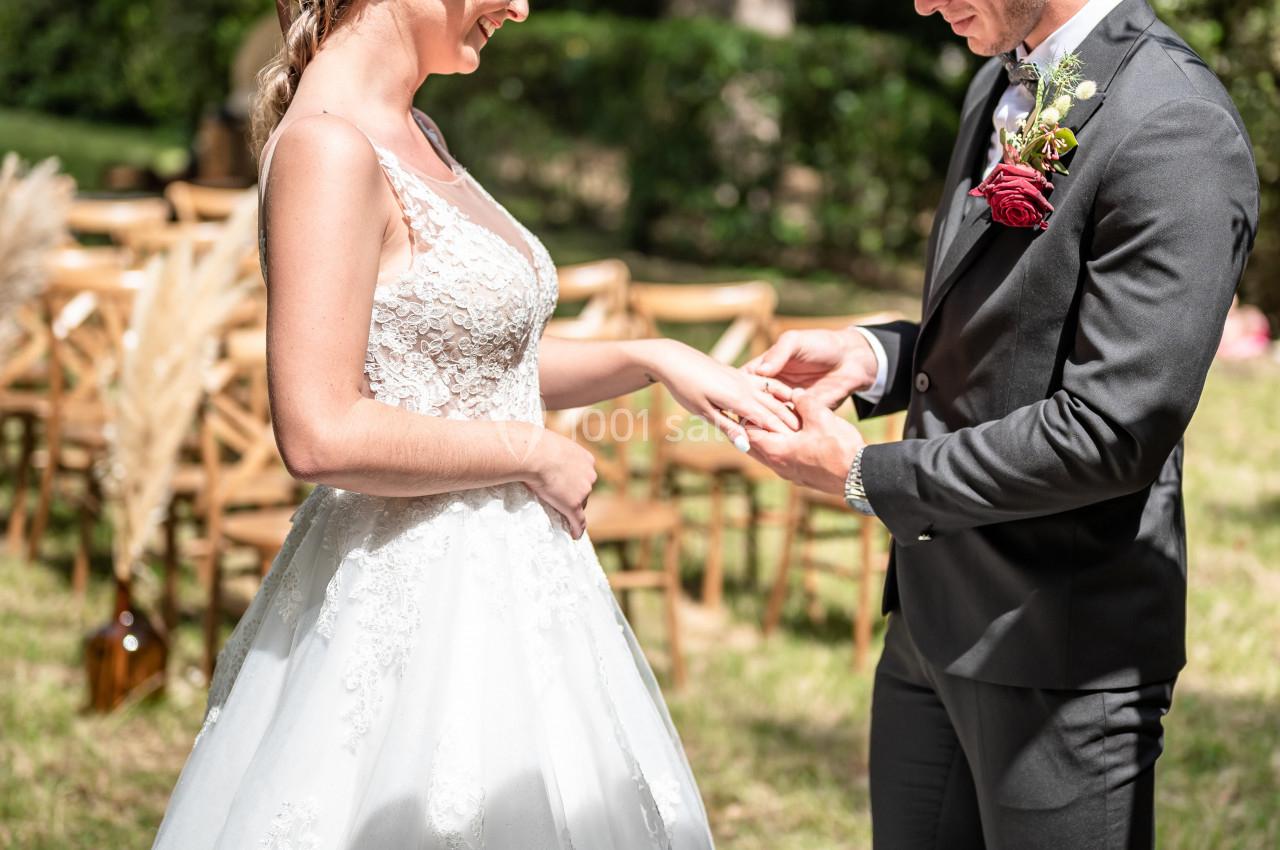 Location salle Malviès (Aude) - Château Guilhem #3 Un couple échange des alliances lors d'une cérémonie de mariage en extérieur, entouré de verdure et de chaises en bois.