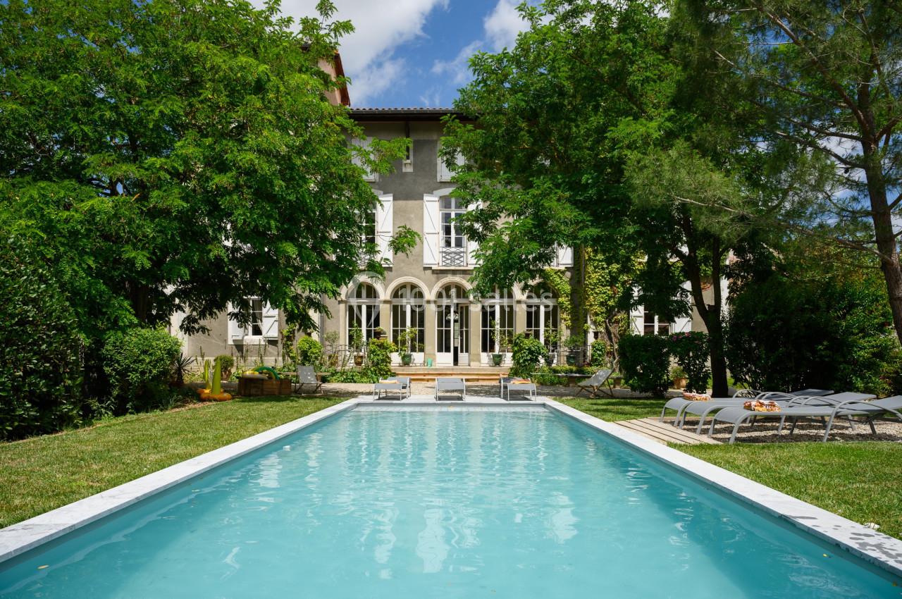 CHATEAU DE STYLE DIRECTOIRE Grande maison entourée d'arbres avec une piscine rectangulaire au premier plan et des chaises longues sur le côté.