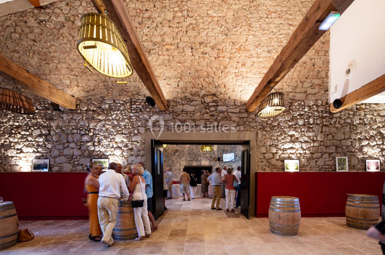 COVENTIS DONNANT SUR COMEDIS Salle en pierre avec poutres apparentes, visiteurs debout près de tonneaux et comptoirs rouges, éclairage chaleureux.