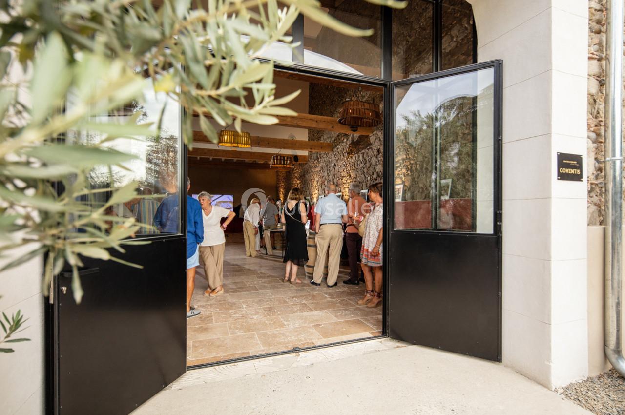 ENTREE COVENTIS Entrée ouverte d'un bâtiment en pierre, avec des personnes rassemblées à l'intérieur dans un espace lumineux.
