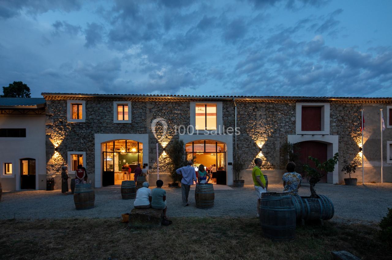EXTERIEUR Façade en pierre d'un bâtiment éclairé au crépuscule, avec des personnes assises et debout devant des tonneaux.