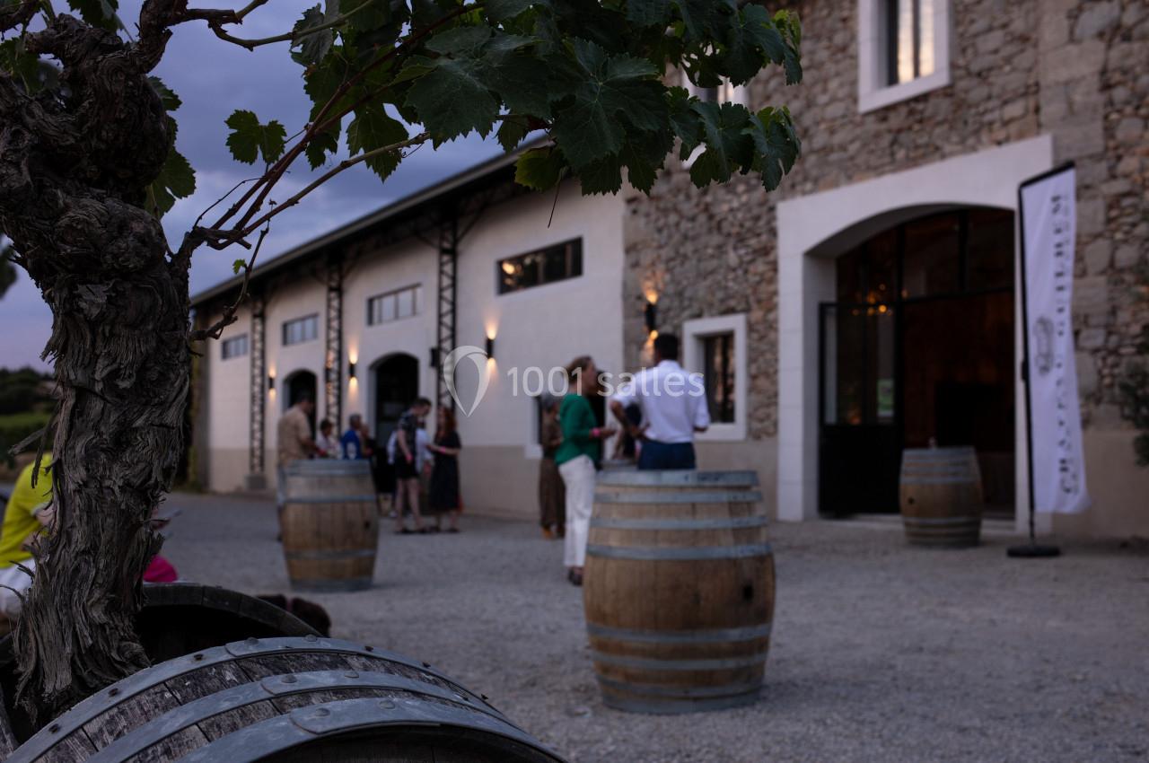 EXTERIEUR Des personnes discutent près de tonneaux en bois devant un bâtiment en pierre, dans une ambiance de fin de journée.